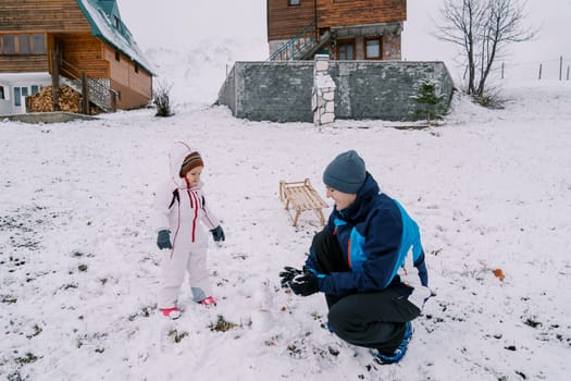 Little girl looks at her dad making a snowball on his haunches near a wooden house. High quality photo