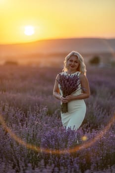 Blonde woman poses in lavender field at sunset. Happy woman in white dress holds lavender bouquet. Aromatherapy concept, lavender oil, photo session in lavender.