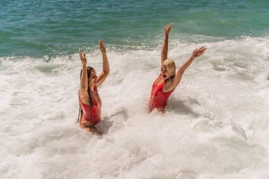 Women ocean play. Seaside, beach daytime, enjoying beach fun. Two women in red swimsuits enjoying themselves in the ocean waves and raising their hands up