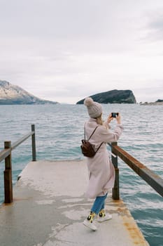 Girl stands on the pier and takes pictures of the sea and mountains with a smartphone. Back view. High quality photo