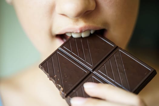 Closeup of crop unrecognizable young girl biting delicious chocolate bar at home