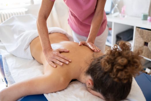 High angle of crop anonymous female osteopath in pink uniform massaging back of woman lying on couch during recovery process