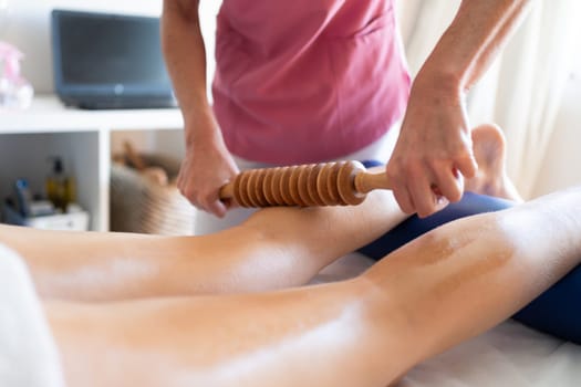 Crop anonymous female osteopathy therapist using wooden device while massaging leg of patient during physiotherapy session in bright modern clinic