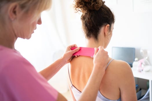 Crop focused professional osteopath applying kinesiology tape on back of neck of unrecognizable young female patient during physiotherapy session in clinic