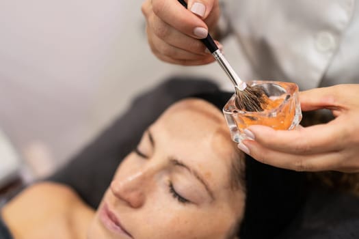 From above of anonymous female beautician looking down, while standing near lady client lying with eyes closed on bed and preparing skin treatment moisturizing product with brush in beauty salon