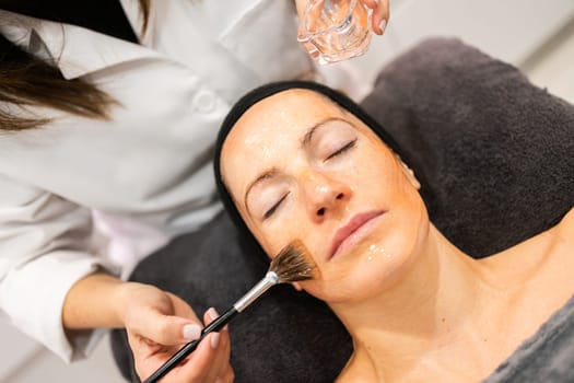 From above of crop anonymous beautician applying cosmetic skincare product with brush on face of woman in beauty salon