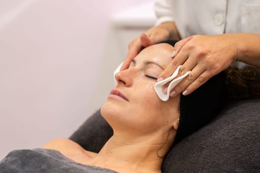 Crop anonymous cosmetician removing skin care product from face of female client with cotton pads during professional facial treatment
