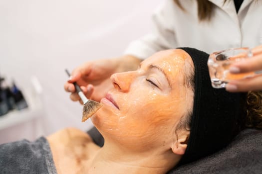 Crop professional beautician applying liquid cosmetic gel mask on face of female client with brush during skin care treatment in beauty salon