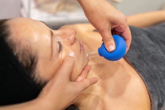 From above view of dark haired woman in gray towel lying and receiving facial cupping massage from anonymous masseuse during skin treatment in beauty center