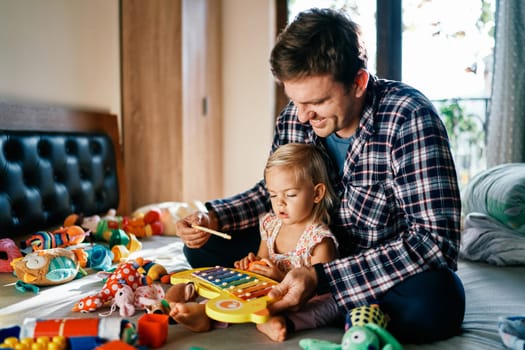 Dad plays with a little girl on a colorful xylophone, sitting behind her on the bed. High quality photo