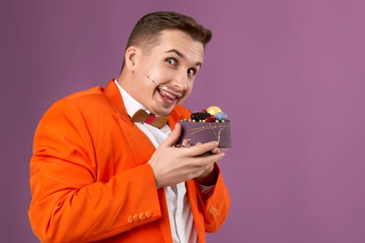 handsome birthday man in orange jacket eating cake on purple background. copy space