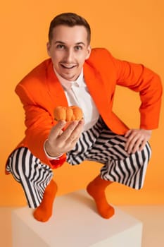 extravagant funny young man in orange jacket and striped pants holding cupcake on orange background.
