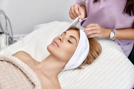 Cosmetologist putting white headband towel on female patient head in beauty salon