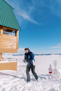 Small child stands near a half-finished snowman while dad walks towards a wooden house, dusting off his mittens. High quality photo