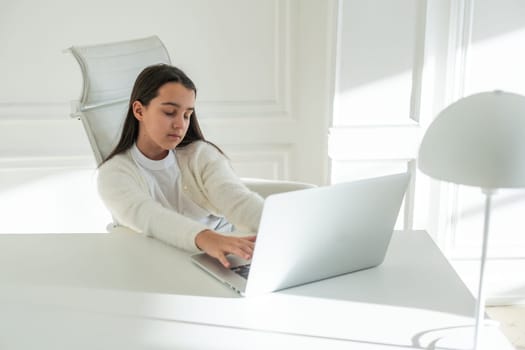 A beautiful sad girl is sitting with a laptop in the kitchen. A serious student doing her homework, working on a research project on the Internet, doing her homework. Online education, home teaching. High quality photo