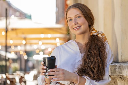 Happy redhead teenager girl enjoying morning coffee hot drink and smiling. Relaxing, taking a break. Young woman standing in urban city center street, drinking coffee to go. Town lifestyles outside