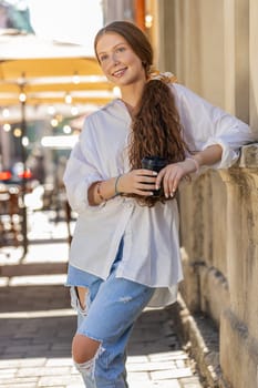 Happy redhead teenager girl enjoying morning coffee hot drink and smiling. Relaxing, taking a break. Young woman standing in urban city center street, drinking coffee to go. Town outside. Vertical