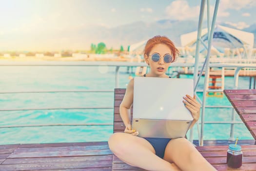 Redhead girl working with a laptop on the sea. Female freelancer on the beach.