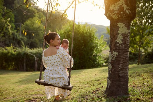 Rear view of a multiracial woman, young adult mother holding her newborn baby while sitting on the swing in the park at sunset, dreamily looking into the distance. Babyhood and maternity leave concept