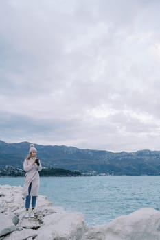 Girl with a phone stands on a rocky seashore against the backdrop of mountains. High quality photo