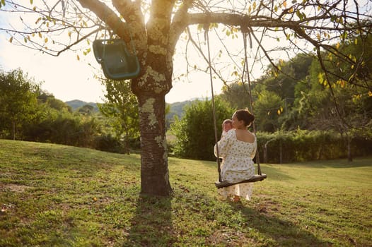 View from the back to a happy young mother holding and kissing her newborn baby, swinging slowly together on the swing in the garden at sunset. People. Babyhood. Motherhood. Maternity leave lifestyle