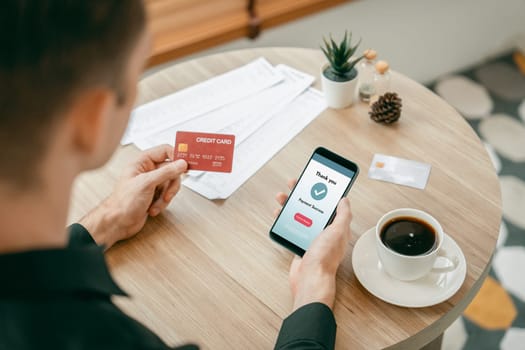 Young man sitting at table using online payment app and digital wallet on smartphone to pay with credit card. E commerce shopping and modern purchasing via mobile internet. Unveiling