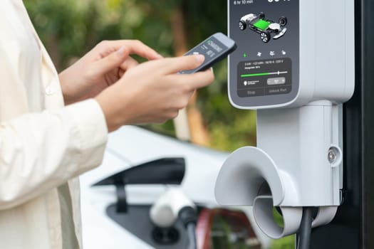 Young woman use smartphone to pay for electricity at public EV car charging station green city park. Modern environmental and sustainable urban lifestyle with EV vehicle. Expedient