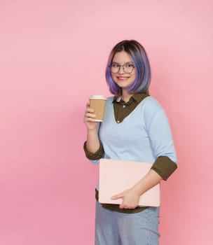 Smiling pupil girl teenager, wearing glasses, immersed in studies with folded laptop and cup of coffee or tea on pink background. Student showcases joyful expression, embodying essence of modern teenage life. High quality photo