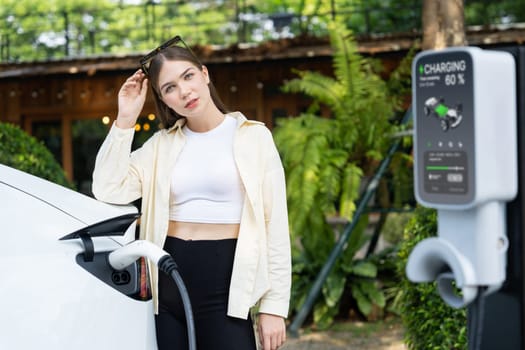 Young woman and sustainable urban commute with EV electric car recharging at outdoor cafe in springtime garden, green city sustainability and environmental friendly EV car. Expedient