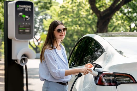 Young woman recharging battery for electric car during road trip travel EV car in natural forest or national park. Eco friendly travel during vacation and holiday. Exalt