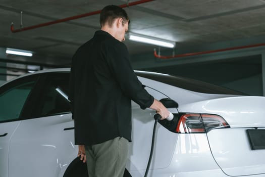 Young man travel with EV electric car to shopping center parking lot charging in downtown city showing urban sustainability lifestyle by green clean rechargeable energy of electric vehicle innards