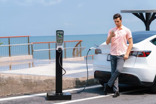 Holiday road trip vacation traveling by the seashore road with electric car, man recharge EV car with green and clean energy while using smartphone. Seascaoe travel with eco-friendly concept.Perpetual