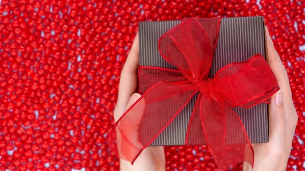 Female's hands holding Christmas gift box decorated with red ribbon bow on red candy background. Christmas and New Year banner. Christmas shopping, Boxing day, pepermint candies