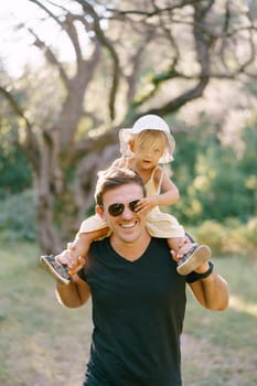 Little daughter grabs sunglasses on dad face while sitting on his shoulders. High quality photo
