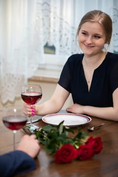 Smiling young blonde woman sitting at the dining room table. She is holding a glass of red wine and looking at the person sitting across from her. There is a bouquet of red roses on the table.