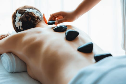 Hot stone massage at spa salon in luxury resort with day light serenity ambient, blissful woman customer enjoying spa basalt stone massage glide over body with soothing warmth. Quiescent