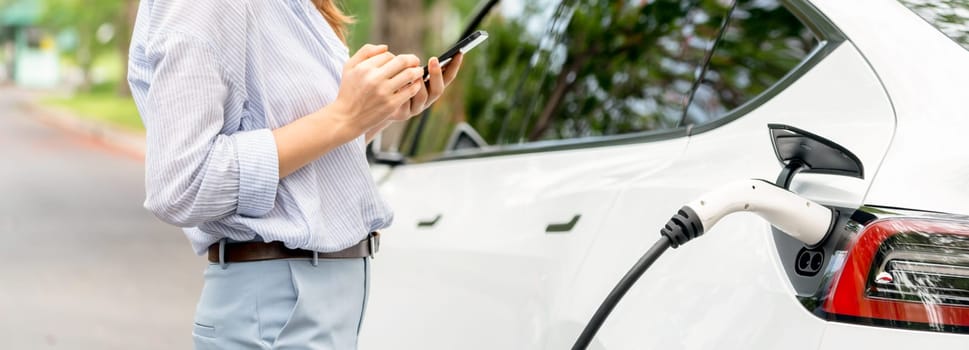 Panorama young woman recharge EV electric vehicle battery from EV charging station and using smartphone online banking to pay for electricity in city park. Eco friendly vehicle with EV car. Exalt