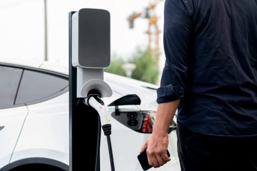 Man using smartphone online banking application to pay for electric car battery charging from EV charging station during vacation holiday road trip at national park or summer forest. Exalt