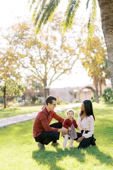 Smiling parents squatting and looking at little girl in sunny park. High quality photo