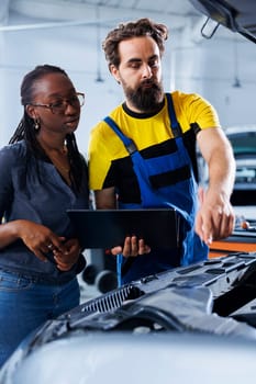 Mechanic in car service uses laptop to calculate invoice after fixing broken automobile distributor. Certified auto repair shop worker uses device to inform woman of final costs after mending vehicle