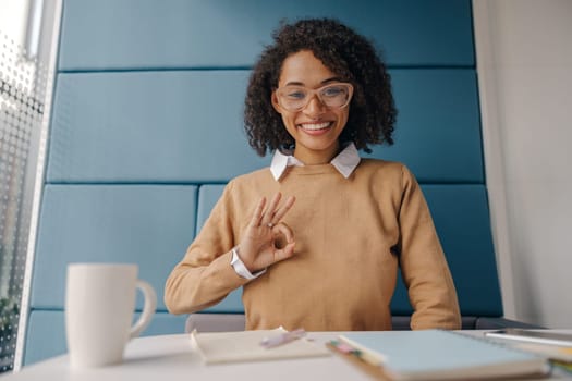 Female freelancer looks camera with smile and showing sign Ok while sitting in cozy coworking