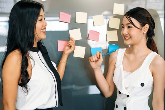 Two young beautiful Asian businesswomen talk and discuss creative post on the window. It is project reminder and thinking idea notes. uds