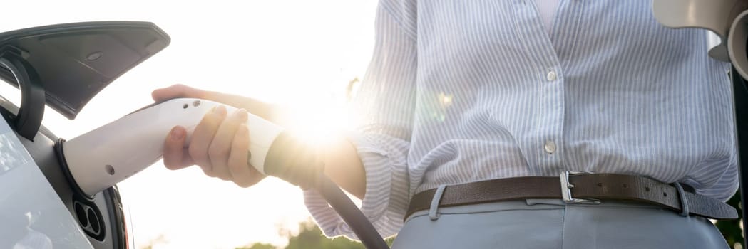 Young woman recharging battery for electric car during road trip travel EV car in natural forest or national park with sunrise lighting. Eco friendly travel during vacation and holiday. Panorama Exalt