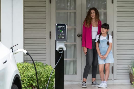 Happy little young girl learn about eco-friendly and energy sustainability as she help her mother recharge electric vehicle from home EV charging station. EV car and modern family. Panorama Synchronos