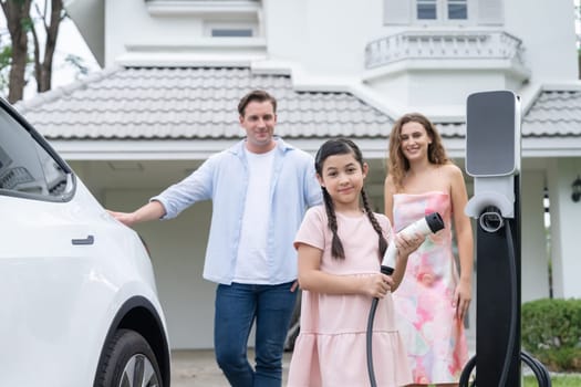 Happy little young girl learn about eco-friendly and energy sustainability as she help her family recharge electric vehicle from home EV charging station. EV car and modern family concept. Synchronos