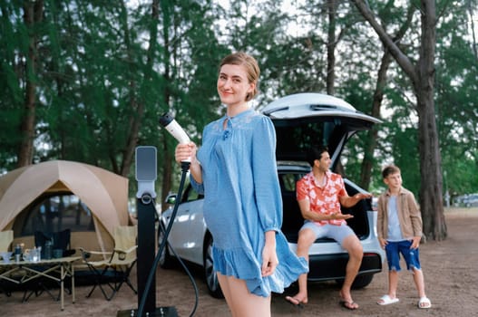 Outdoor adventure and family vacation camping at sea travel by eco friendly car. Cheerful woman or mother holding, pointing EV charger point with playful and happiness posture in campsite. Perpetual