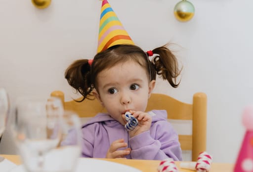 One beautiful Caucasian girl with two ponytails and a cone hat on her head sits on a chair at the festive table and blows a whistle on a paper pipe while celebrating her birthday, close-up side view.