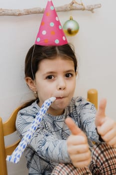 One beautiful Caucasian brunette girl with big eyes and a pink cone hat on her head sits on a chair at the festive table and blows a whistle into a paper pipe and shows a cool gesture with her fingers, celebrating her birthday, close-up side view.