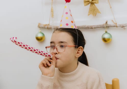 One beautiful Caucasian brunette girl wearing glasses and with a pale pink cone hat on her head sits on a chair at the festive table and blows a whistle into a paper pipe, celebrating her birthday, close-up side view.