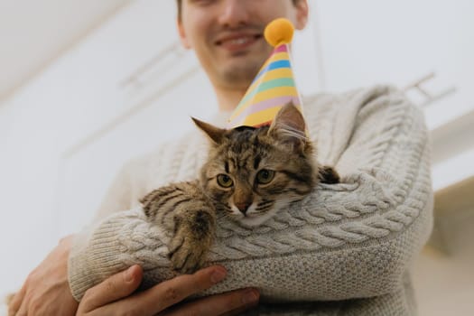 One handsome Caucasian happy guy holding a small tricolor kitten with a cone hat on his head, standing in the kitchen and celebrating a birthday, close-up view from below.